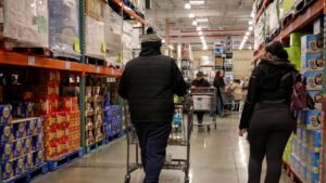 People shopping at Costco, aisles of groceries, bulk items on shelves.